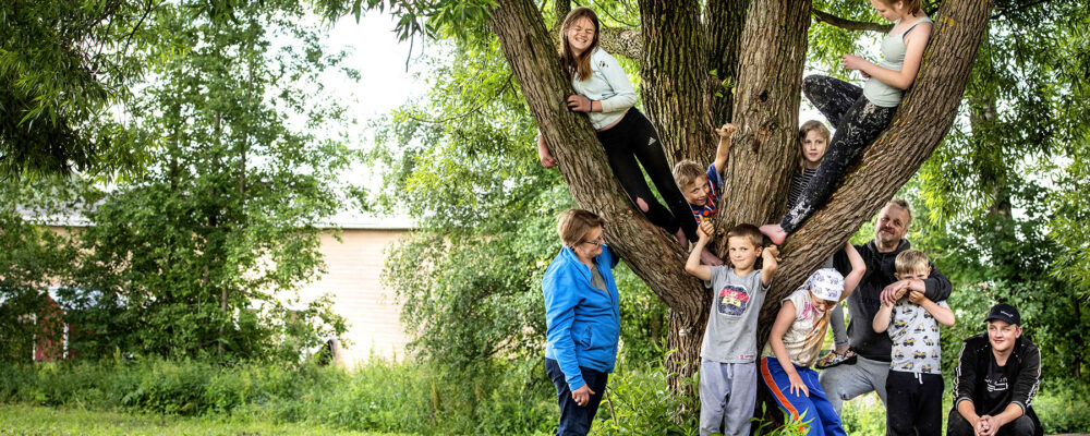 Kaksi aikuista ja iso joukko lapsia kokoontuneena puun ympärille.