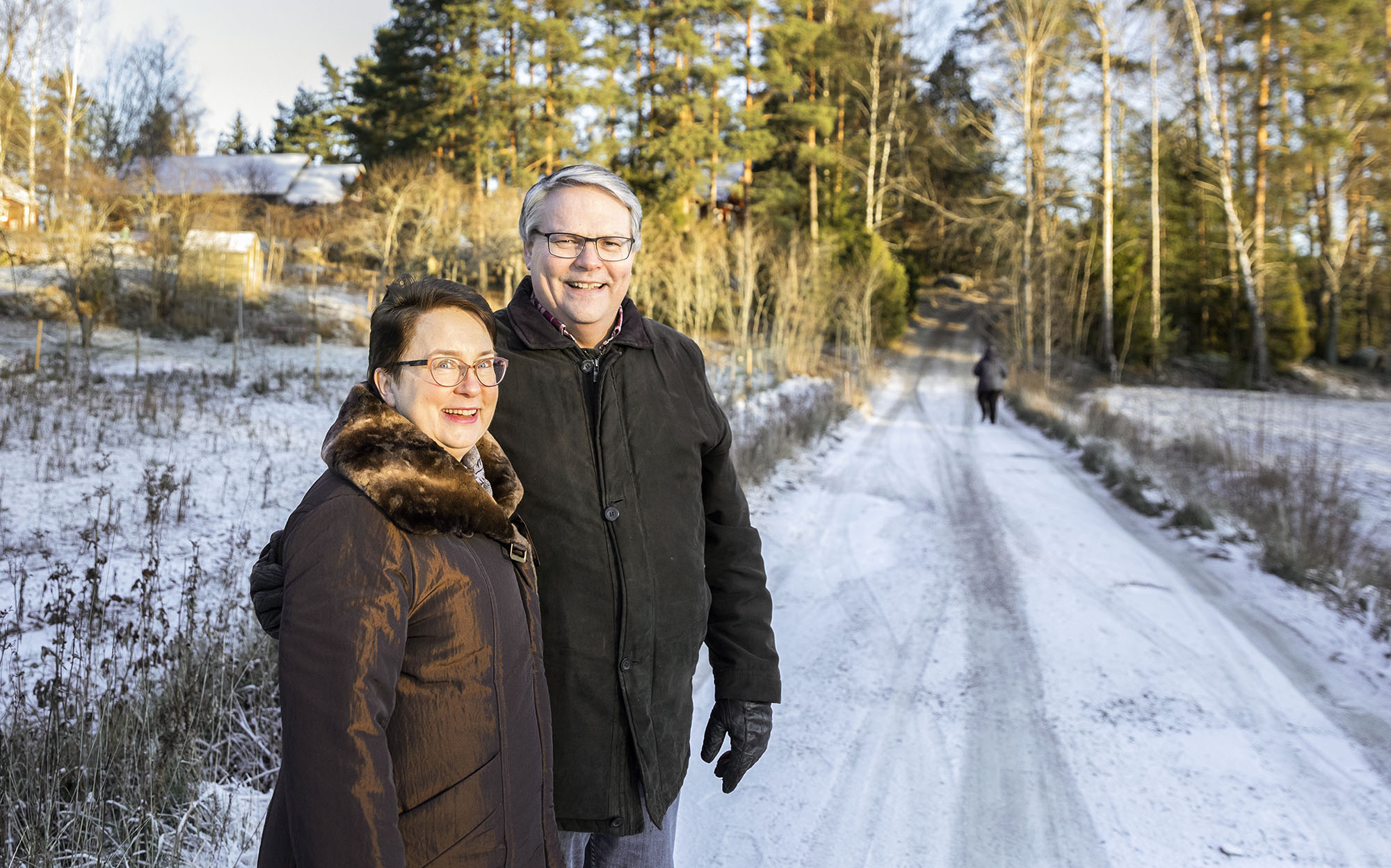 Mies ja nainen seisoo metsätien varressa. Maassa on vähän lunta. Taustalla näkyy taloja, metsikköä ja tietä pitkin kävelevä ihminen.