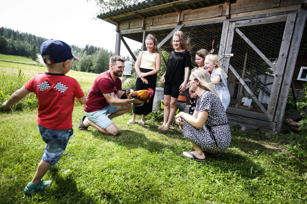 Mies, nainen ja viisi lasta ovat ulkona kanalan edessä. He kaikki katselevat kukkoa, jota mies pitelee käsissään.