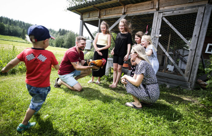 Mies, nainen ja viisi lasta ovat ulkona kanalan edessä. He kaikki katselevat kukkoa, jota mies pitelee käsissään.