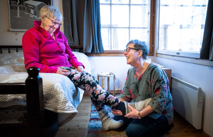 Nainen avustaa vanhempaa naista sukkien jalkaan laittamisessa sängyn laidalla.