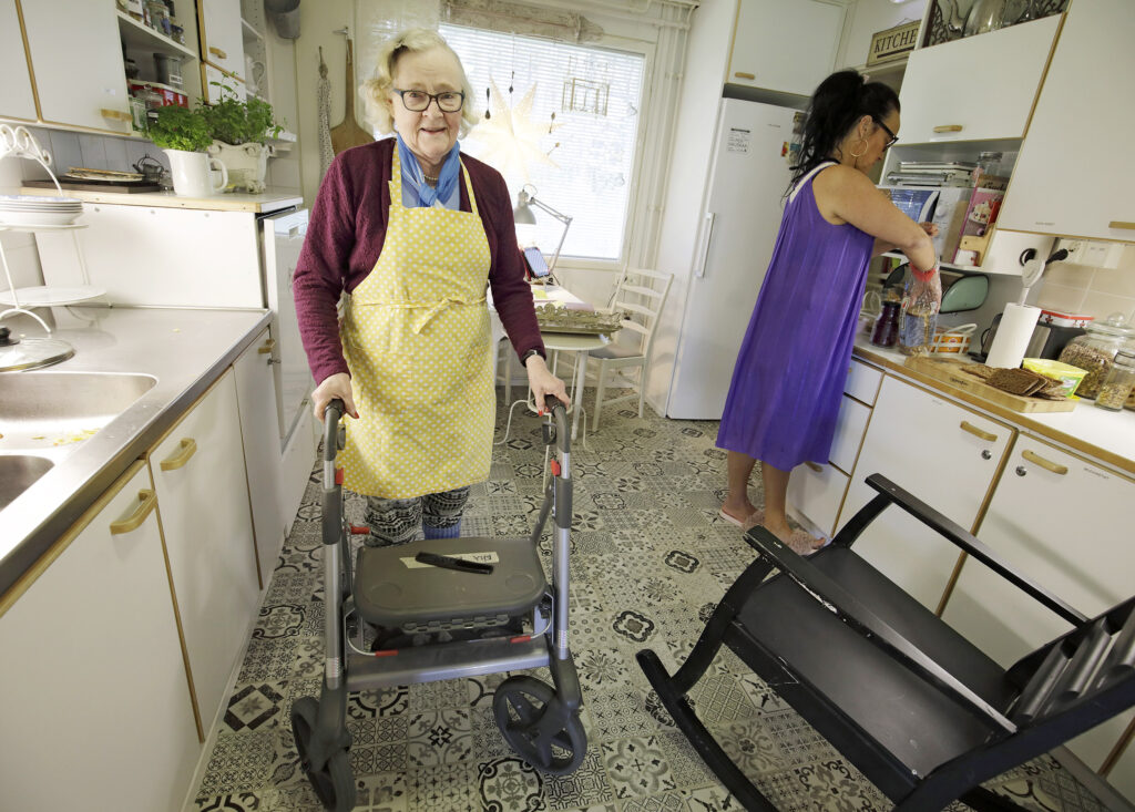 Vanha nainen kävelee keittiössä rollaattorin kanssa. Taustalla nainen laittaa ruokaa.