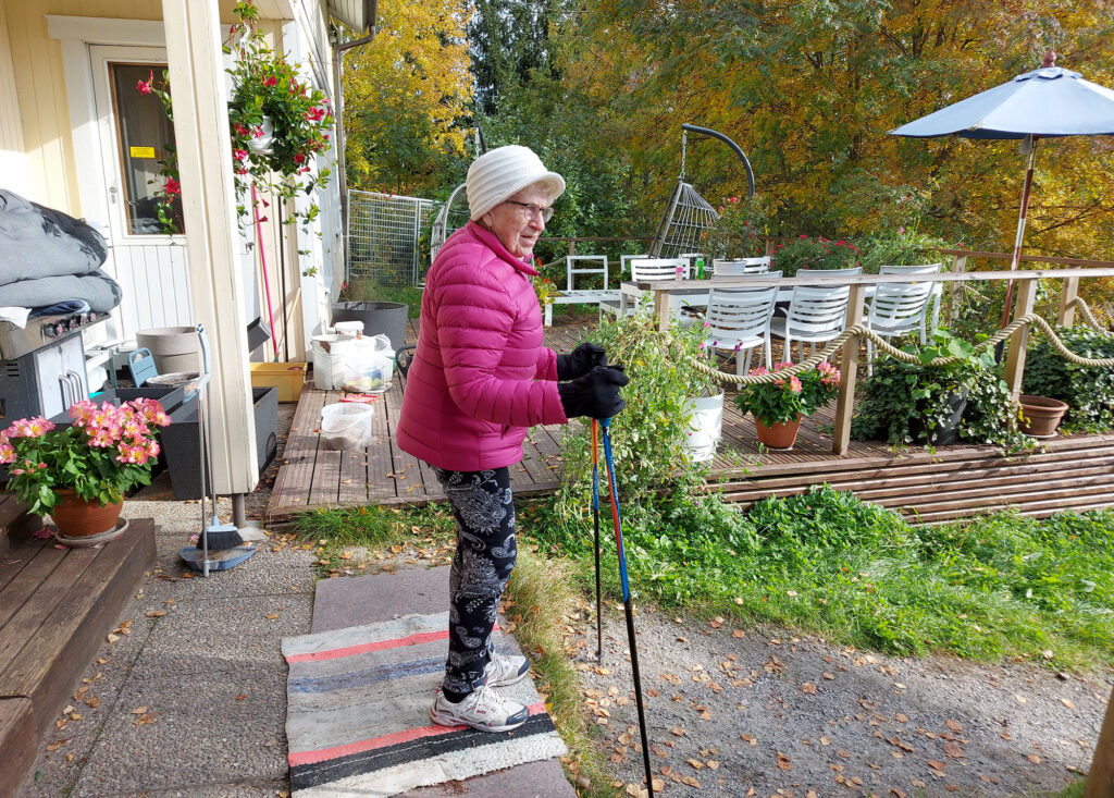 Vanhempi nainen on lähdössä sauvakävelemään omakotitalon pihasta. Taustalla näkyy syksyistä metsää.