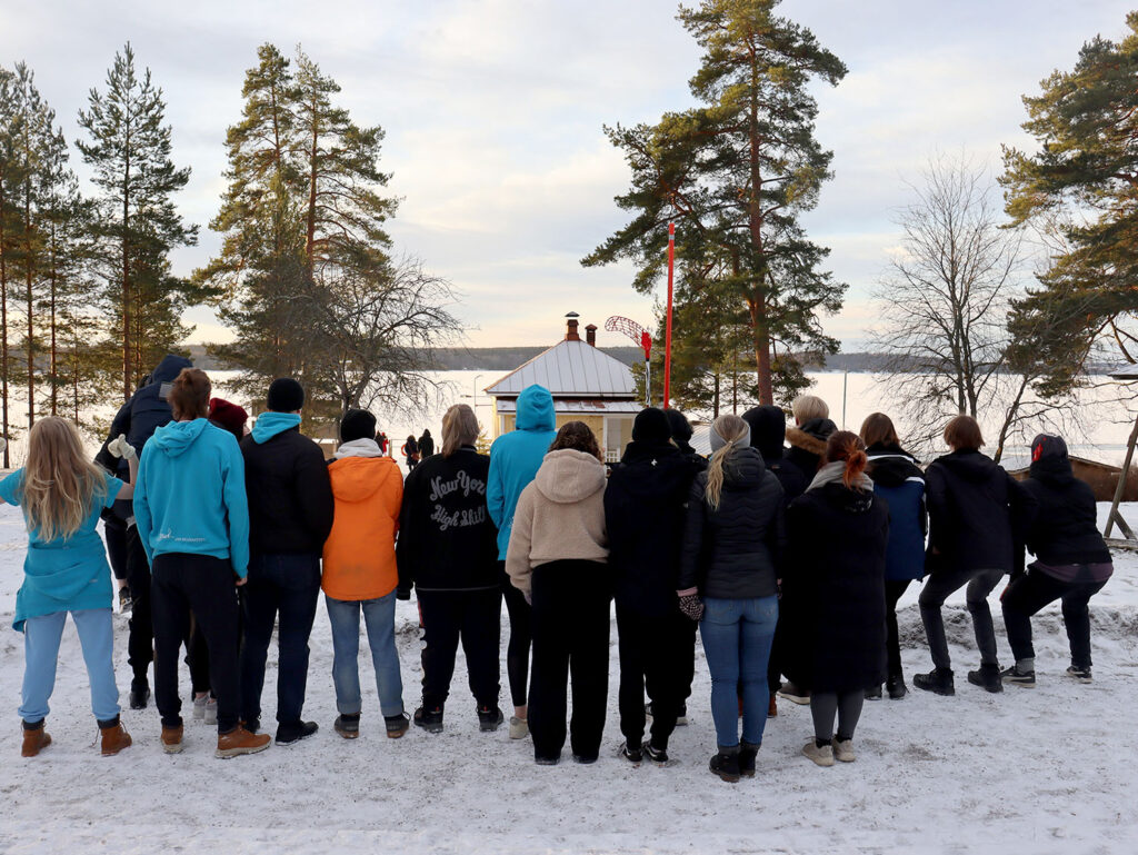 Iso joukko ihmisiä seisoo selkä kameraan päin talvipäivänä järven rannalla.