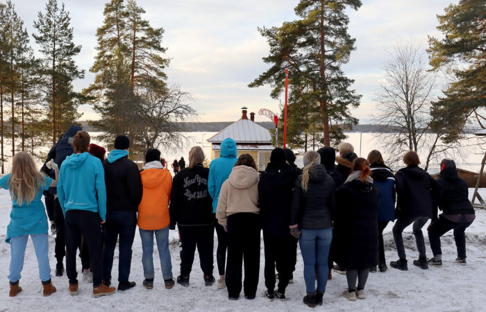 Iso joukko ihmisiä seisoo selkä kameraan päin talvipäivänä järven rannalla.