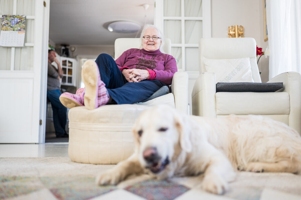 Iäkäs nainen istuu olohuoneen nojatuolilla, hymyilee ja katselee lattialla makaavaa koiraa.