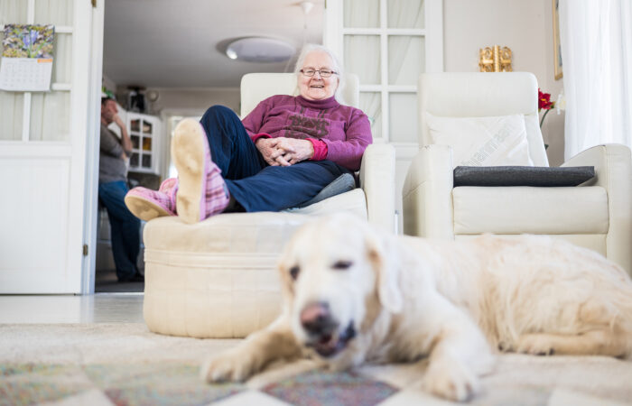 Iäkäs nainen istuu olohuoneen nojatuolilla, hymyilee ja katselee lattialla makaavaa koiraa.