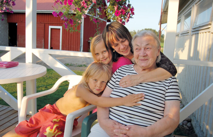 Kaksi lasta, aikuinen ja ikäihminen istuvat maalaistalon kuistilla ja halaavat toisiaan.