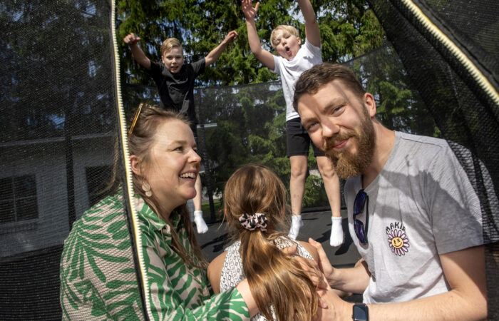 Kaksi lasta pomppii trampoliinilla omakotitalon pihalla, ja muu perhe istuu trampoliinin reunalla.