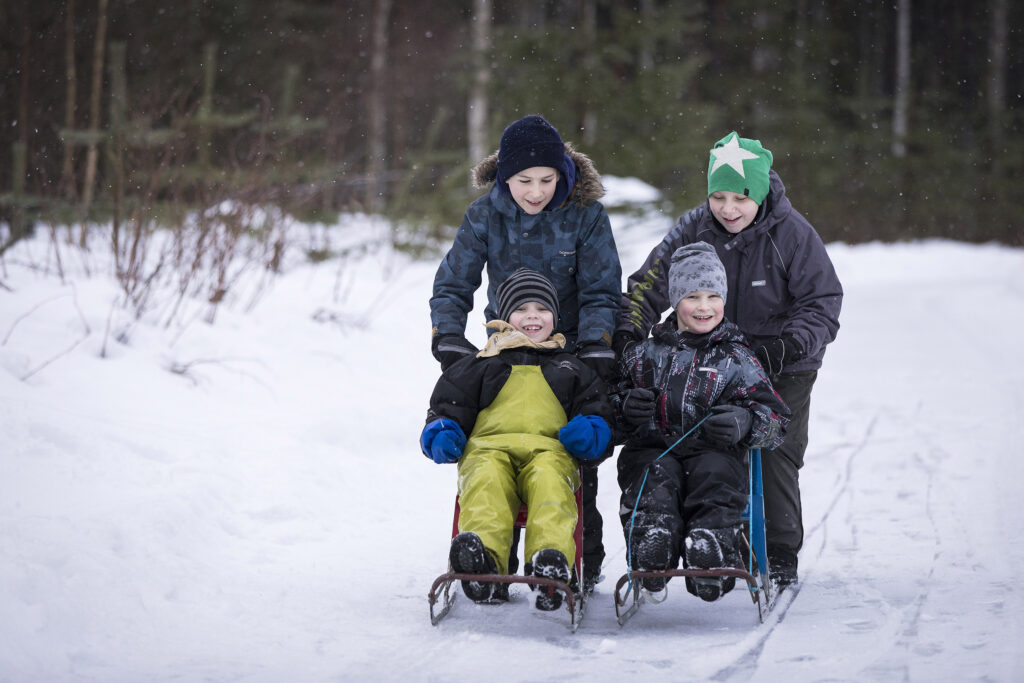 Joukko iloisia lapsia kulkee vauhdikkaasti potkukelkoilla talvisella metsätiellä.