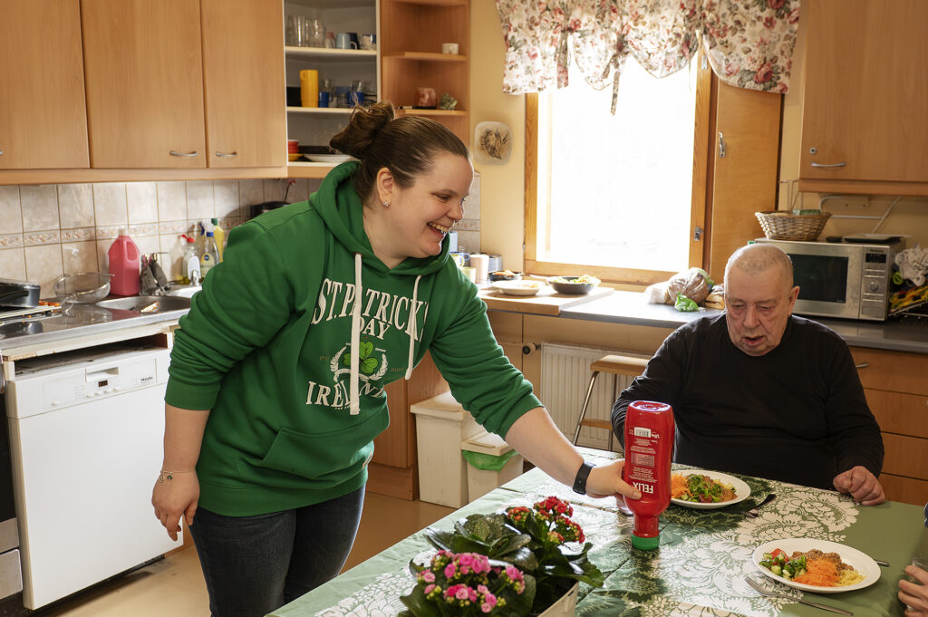 Nainen laittaa ketsuppipulloa ruokapöydälle, jonka ääressä mies istuu ruokalautanen edessään.