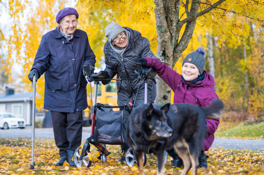 Kaksi iäkästä naista, nuorempi nainen ja koira seisovat keltalehtisen syyspihlajan alla.