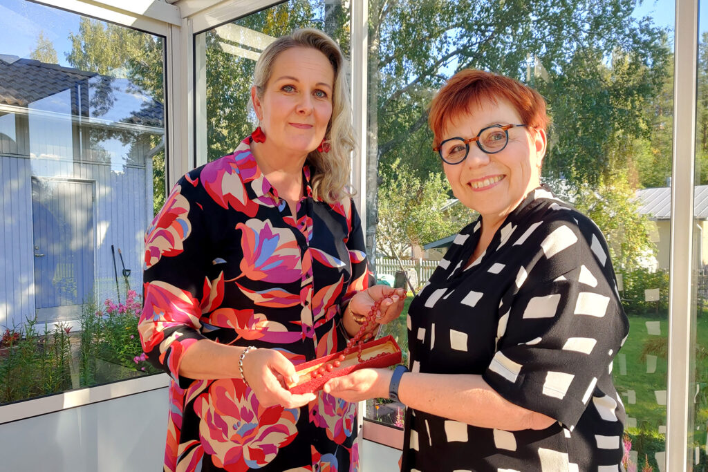 Kaksi naista seisoo lasiverannalla ja pitelee kädessään helminauhaa.
