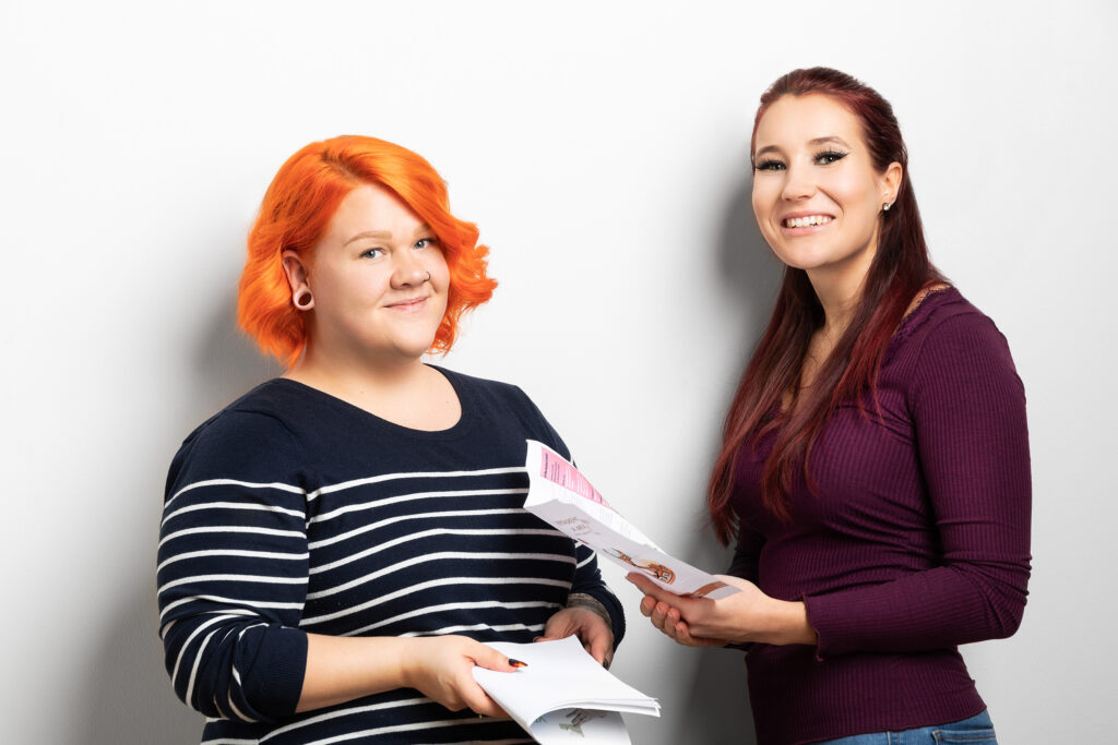 Kaksi nuorta naista naista pitelee paperiesitteitä käsissään ja hymyilee.