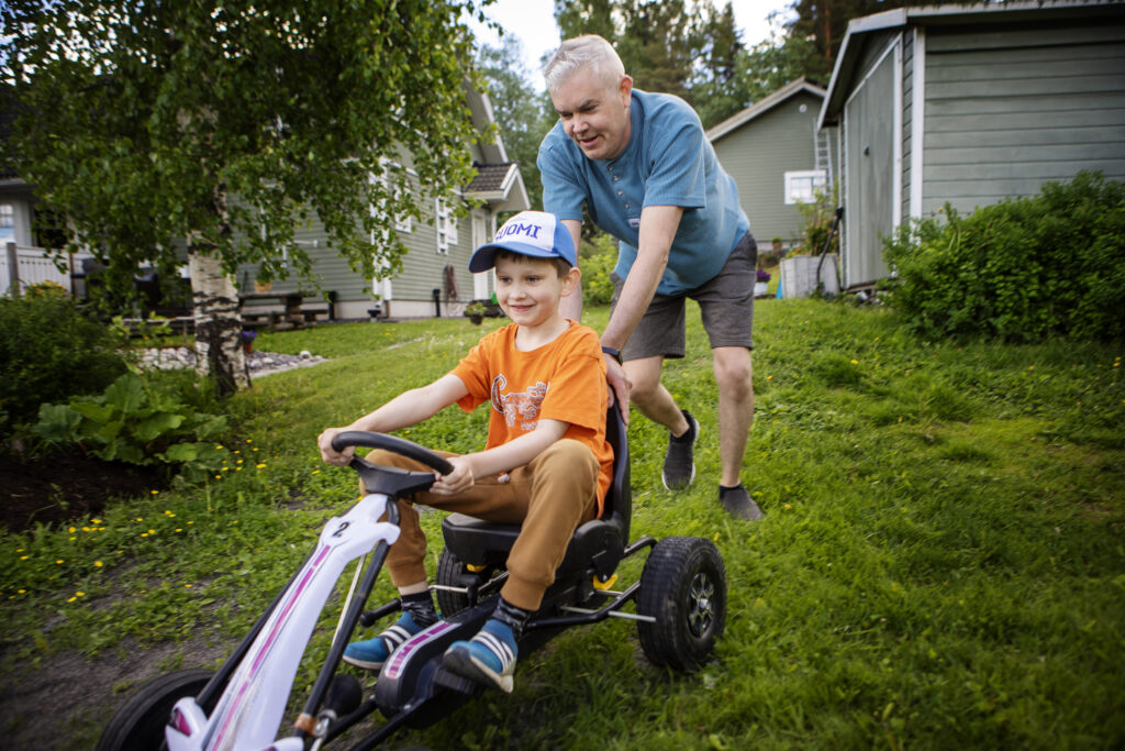 Mies työntää polkuauton kyydissä istuvaa pikkupoikaa omakotitalon nurmikolla.