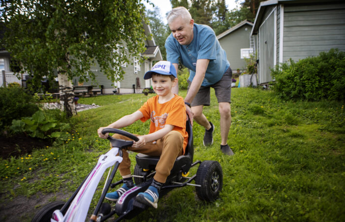 Mies työntää polkuauton kyydissä istuvaa pikkupoikaa omakotitalon nurmikolla.