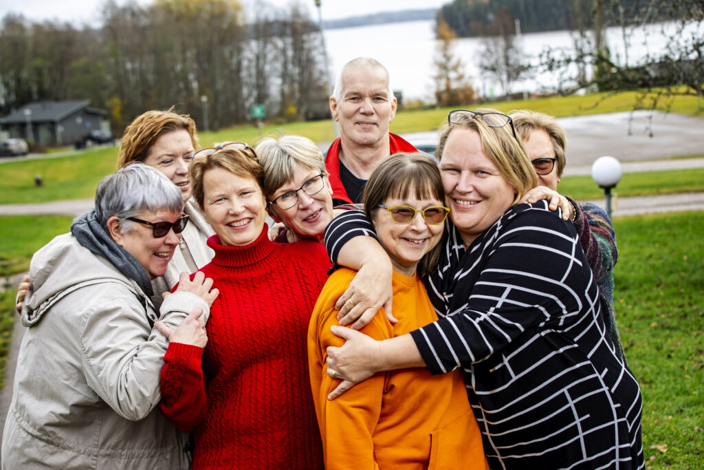 Ryhmä iloisia ihmisiä halaa toisiaan syksyisen järven rantamilla.