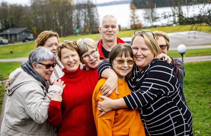 Ryhmä iloisia ihmisiä halaa toisiaan syksyisen järven rantamilla.