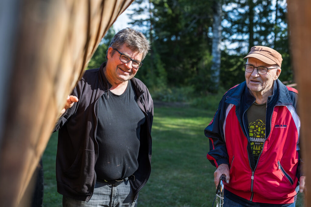 Kaksi miestä katselee nurmikolla olevan puuveneen kylkeä.