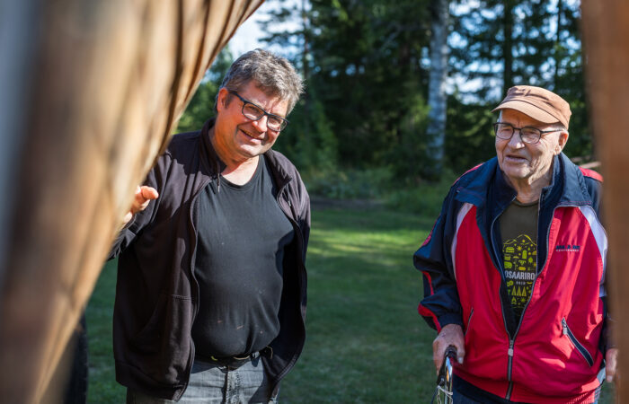 Kaksi miestä katselee nurmikolla olevan puuveneen kylkeä.