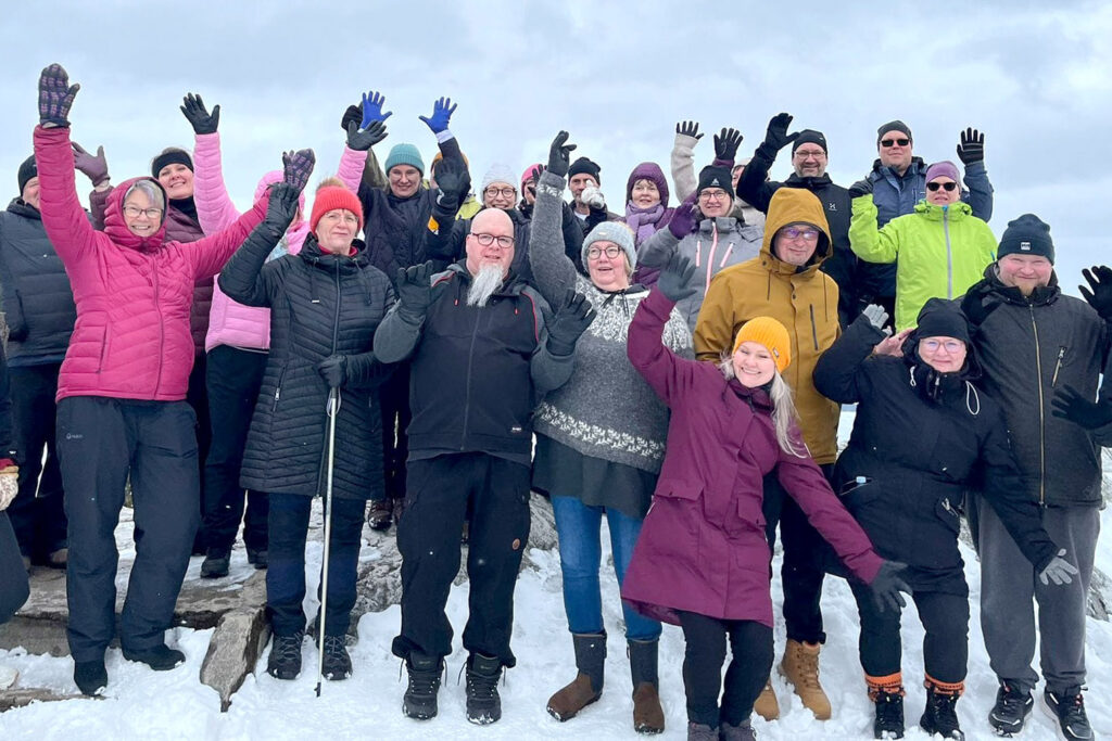 Ryhmä iloisia ihmisiä seisoo lumisella rinteellä ja pitää käsiään pystyssä.