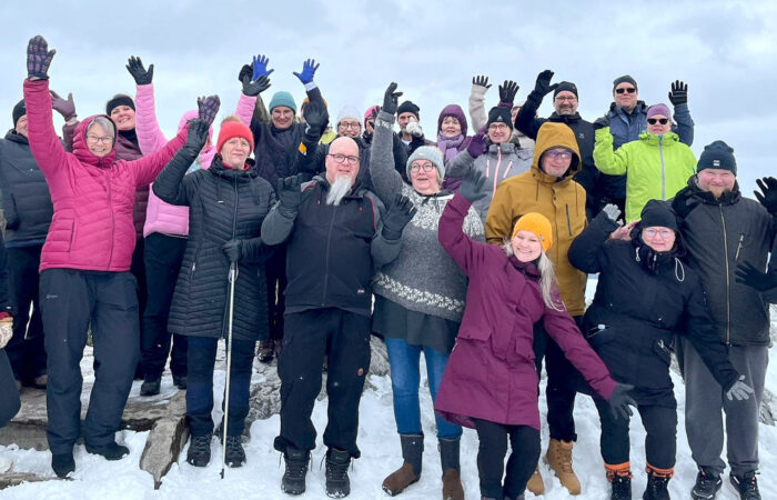 Ryhmä iloisia ihmisiä seisoo lumisella rinteellä ja pitää käsiään pystyssä.
