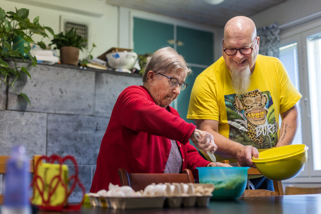 Keltapaitainen mies laittaa ruokaa punapaitaisen iäkkäämmän naisen kanssa keittiössä.