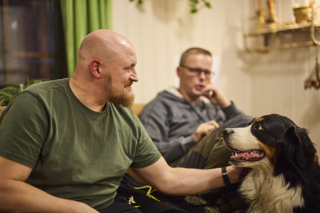 Mies rapsuttelee koiraa olohuoneessa, taustalla toinen mies.