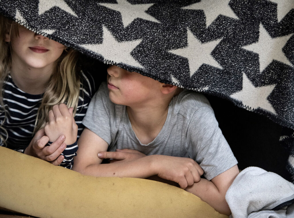 Kaksi lasta makaa sängyllä peitto silmiensä edessä.