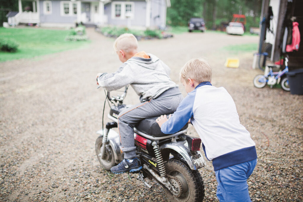 Poika istuu mopon kyydissä omakotitalon pihalla ja toinen poika työntää mopoa.