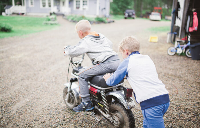 Poika istuu mopon kyydissä omakotitalon pihalla ja toinen poika työntää mopoa.