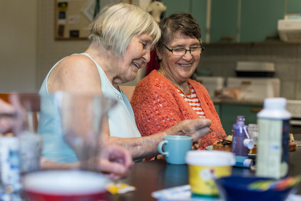 Kaksi naista istuu ruokapöydän ääressä ja leikkaa piirakkaa.