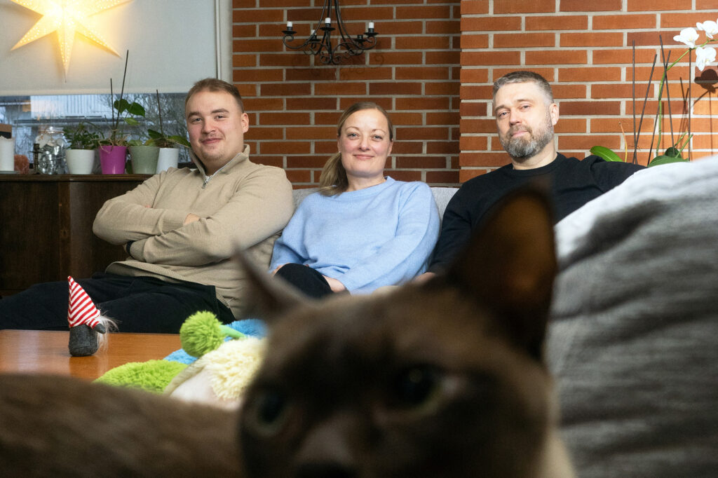 Kaksi miestä ja nainen istuvat sohvalla, etualalla kissa.