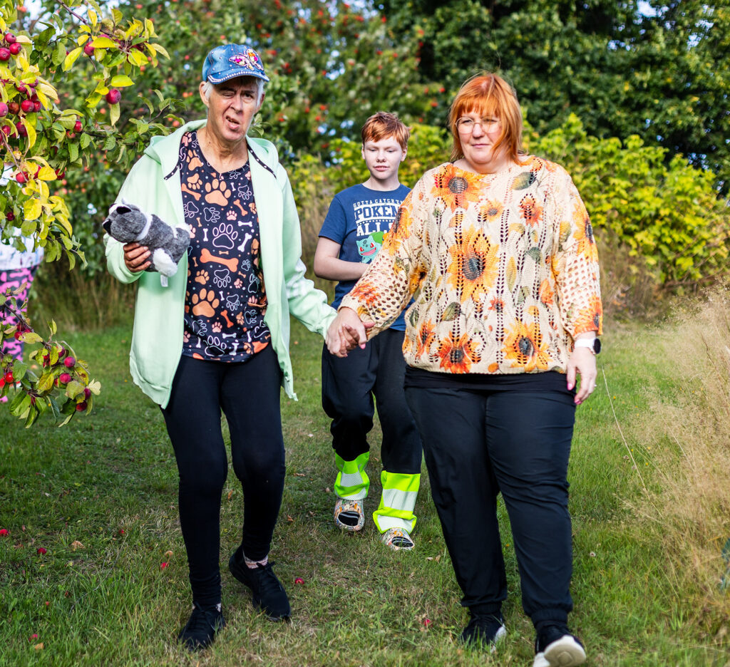 Kaksi aikuista ja lapsi kävelevät nurmikolla omenapuun vieressä.