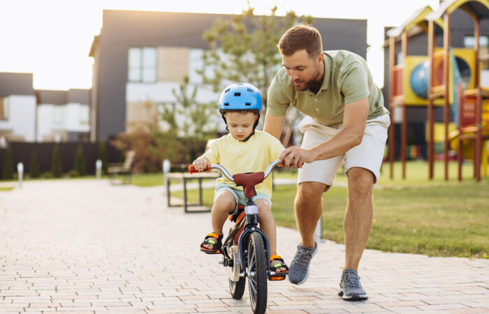 Aikuinen mies opettaa pientä lasta ajamaan polkupyörällä leikkipuiston vieressä.