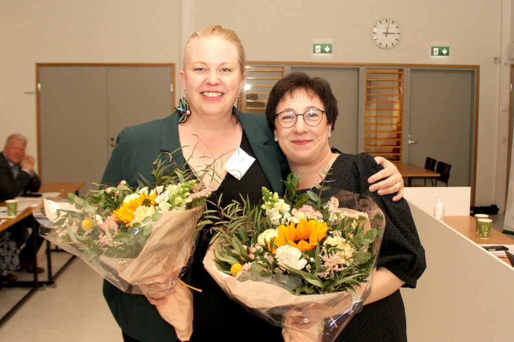 Kaksi naista hymyilee ja halaa toisiaan kukkapaketit sylissään.