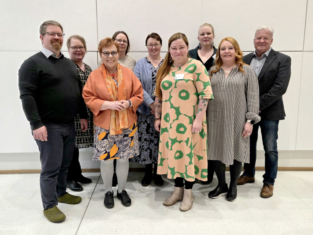 Ryhmä hymyileviä ihmisiä seisoo vaalean seinän edessä.