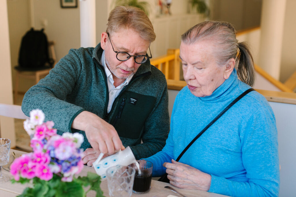 Silmälasipäinen mies kaataa maitoa sinipaitaisen naisen kahvimukiin.
