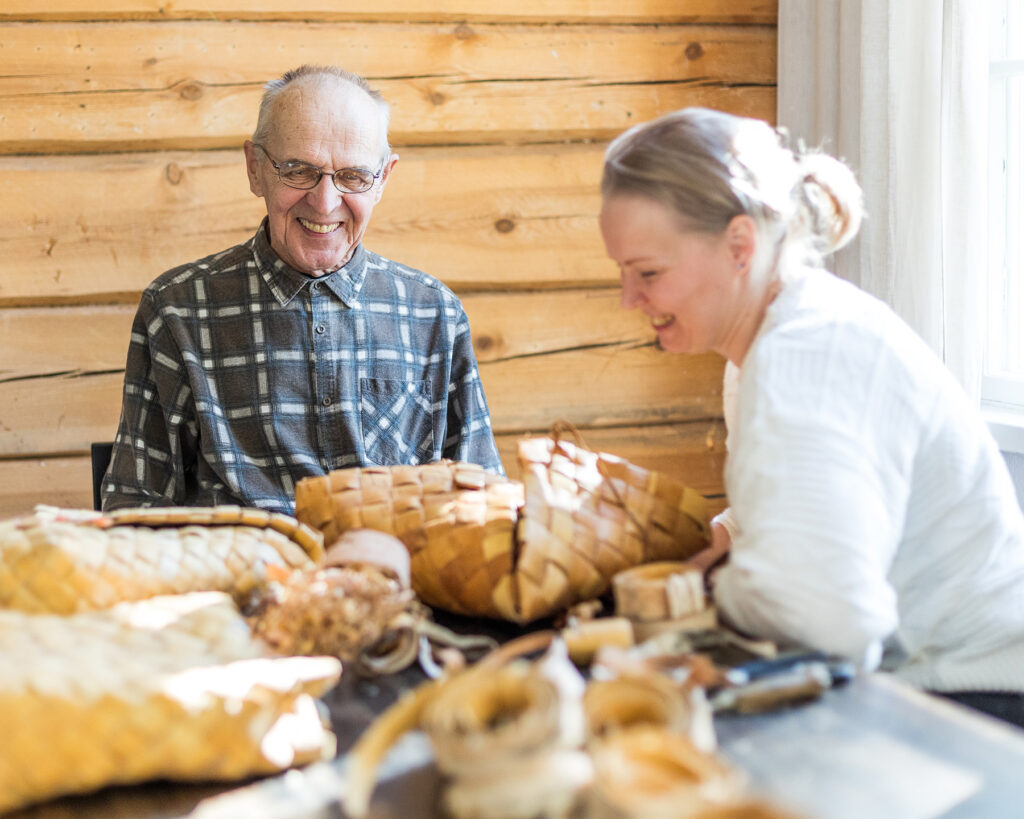 Vanha mies ja nuorempi nainen nauravat pöydän äärellä. Pöydällä on tuohitöitä.