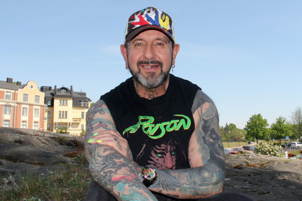 Tatuoitu, lippalakkipäinen mies istuu kalliolla.