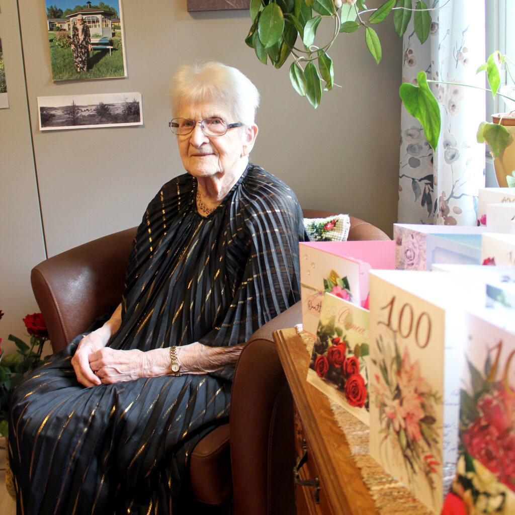 Harmaatukkainen nainen istuu nojatuolilla huoneessa, jossa on onnittelukortteja.